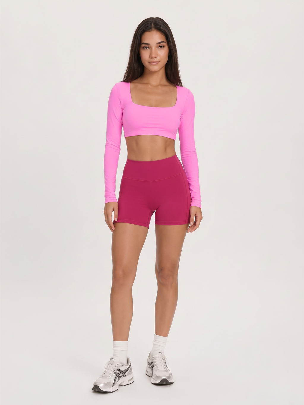 A woman modeling a bright pink long-sleeve crop top and magenta high-waisted shorts against a plain light gray studio background.