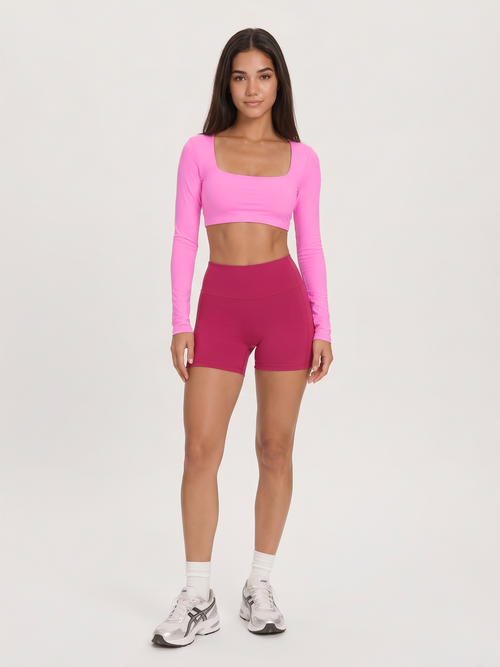A woman modeling a bright pink long-sleeve crop top and magenta high-waisted shorts against a plain light gray studio background.