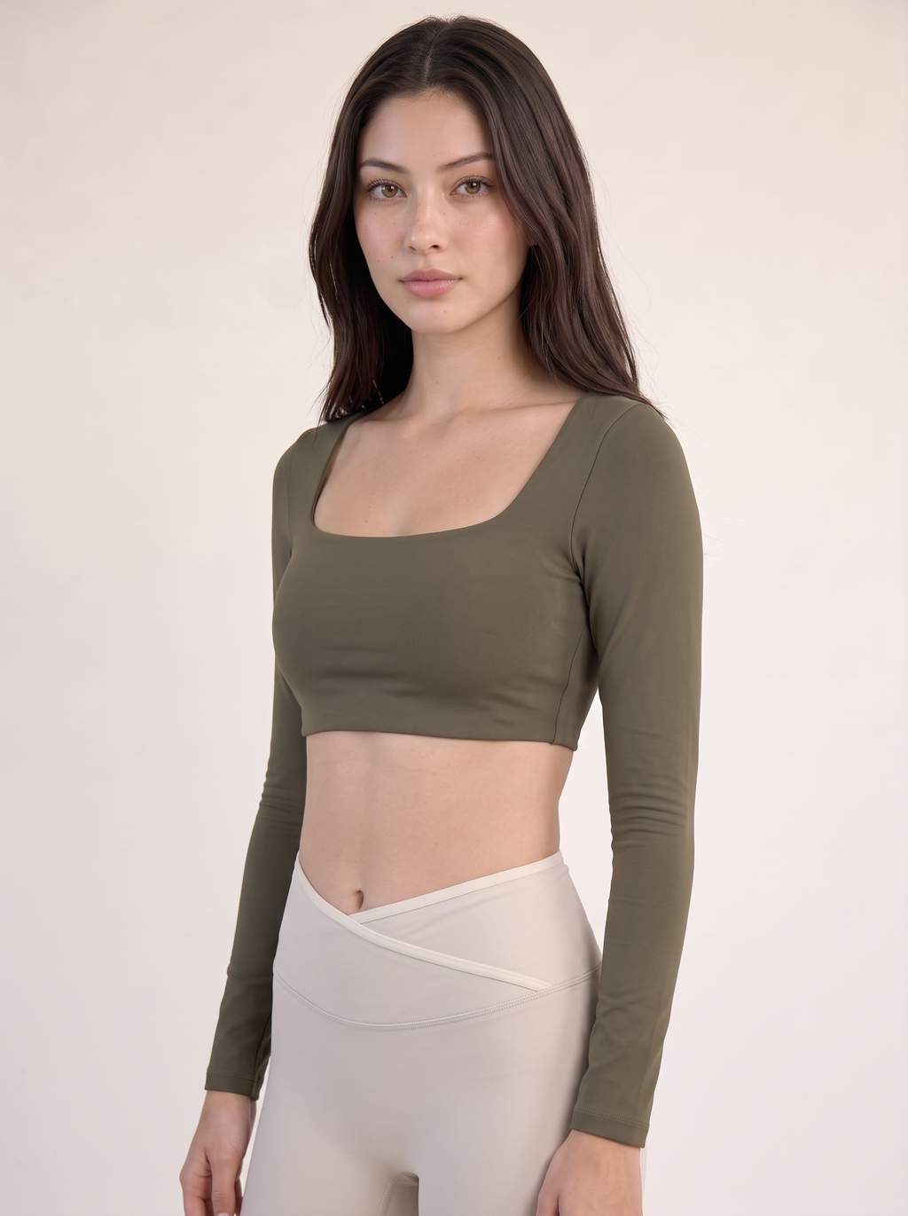 A woman modeling a long-sleeve square-neck crop top in olive green, paired with light beige leggings, against a plain light beige studio background.