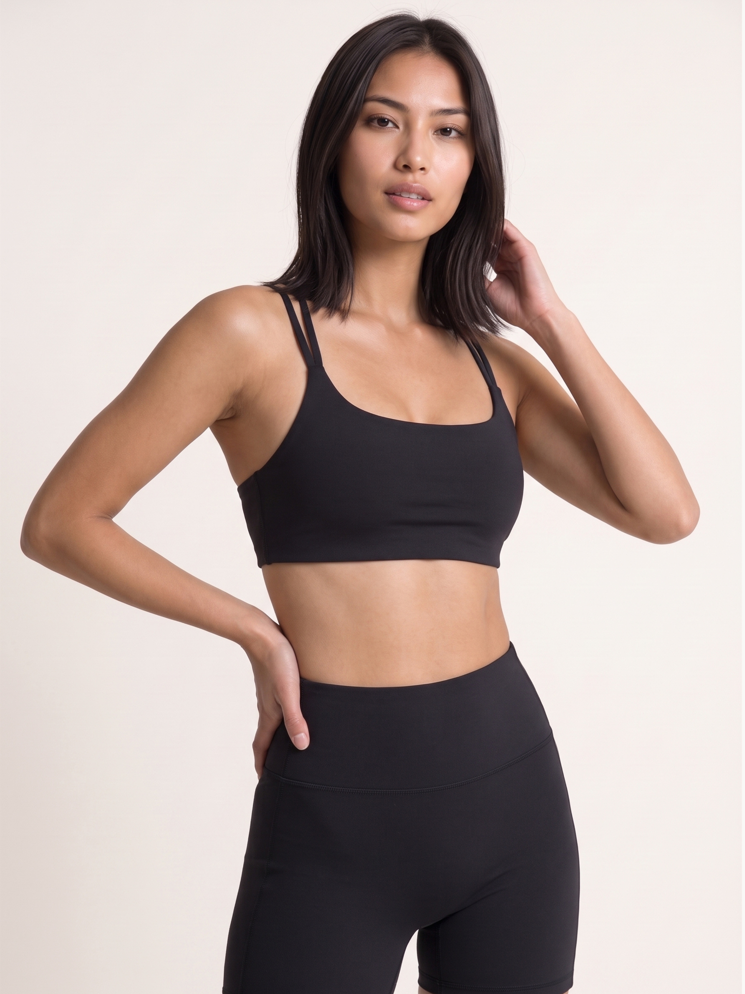 A woman modeling a black sports bra and matching high-waisted black leggings against a plain light beige studio background.