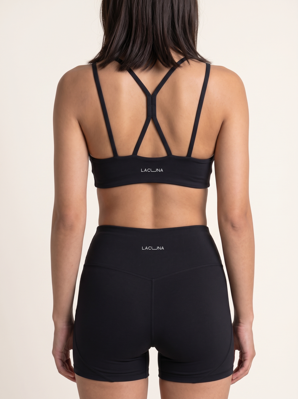 A woman shown from the back wearing a black double straps sports bra and matching high-waisted shorts with the brand name LACUNA printed in white on the waistband and center back.