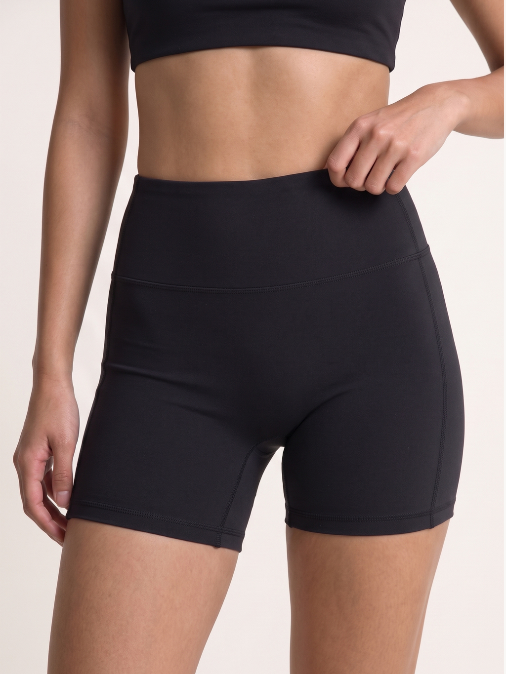 A close-up of a model wearing black high-waisted athletic shorts, showing the waistband and side pocket detail against a light beige background.