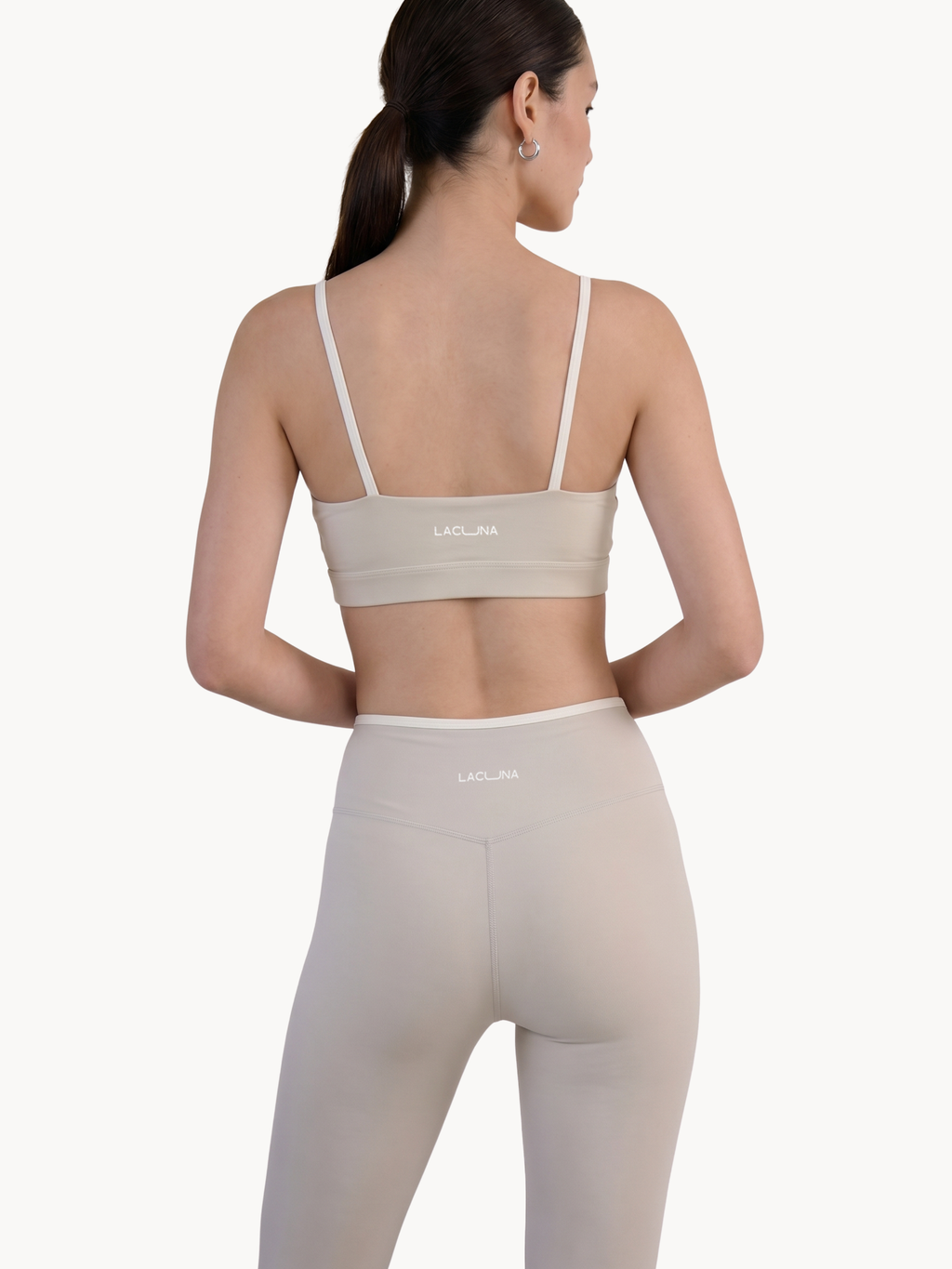 A woman shown from the back wearing a matching beige sports bra and high-waisted leggings set with a small white LACUNA logo on the waistband and back of the bra. Earl Grey