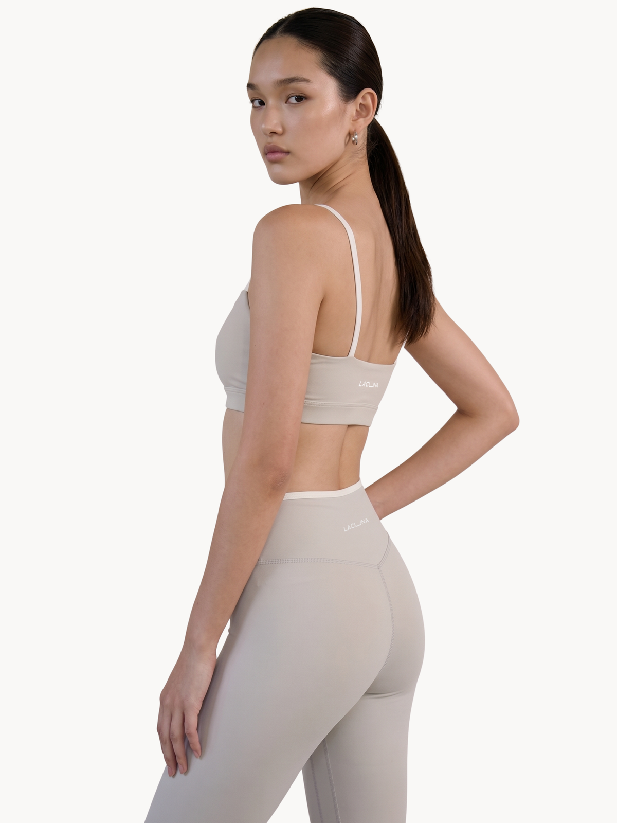 A woman modeling a light beige sports bra and matching high-waisted leggings against a plain white studio background. Earl Grey