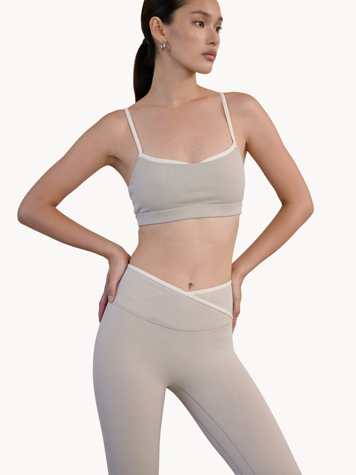A woman modeling a light beige activewear set consisting of a sports bra and high-waisted leggings with contrasting white trim, set against a plain off-white background. Earl Grey