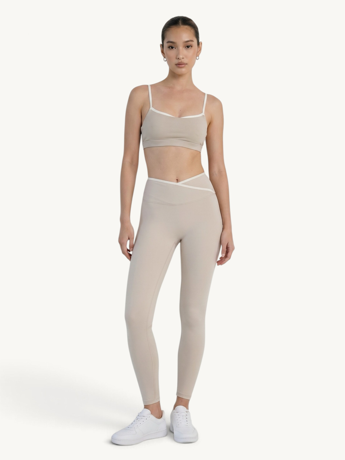 A model wearing a matching beige activewear set consisting of a sports bra and high-waisted leggings with white trim, paired with white sneakers, against a plain off-white background.